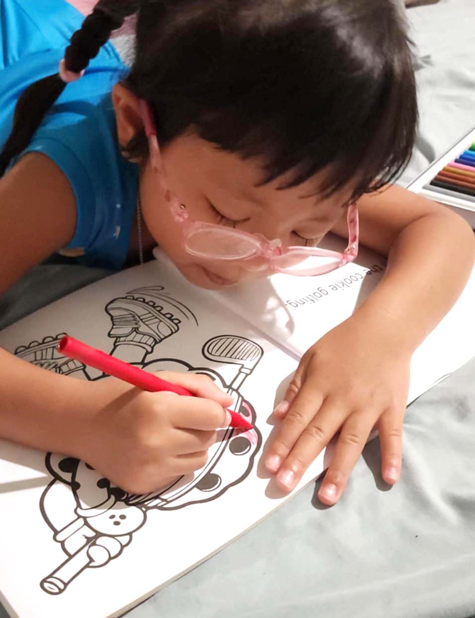 Female child wearing glasses coloring in an Active Snacks coloring book with a red crayon.