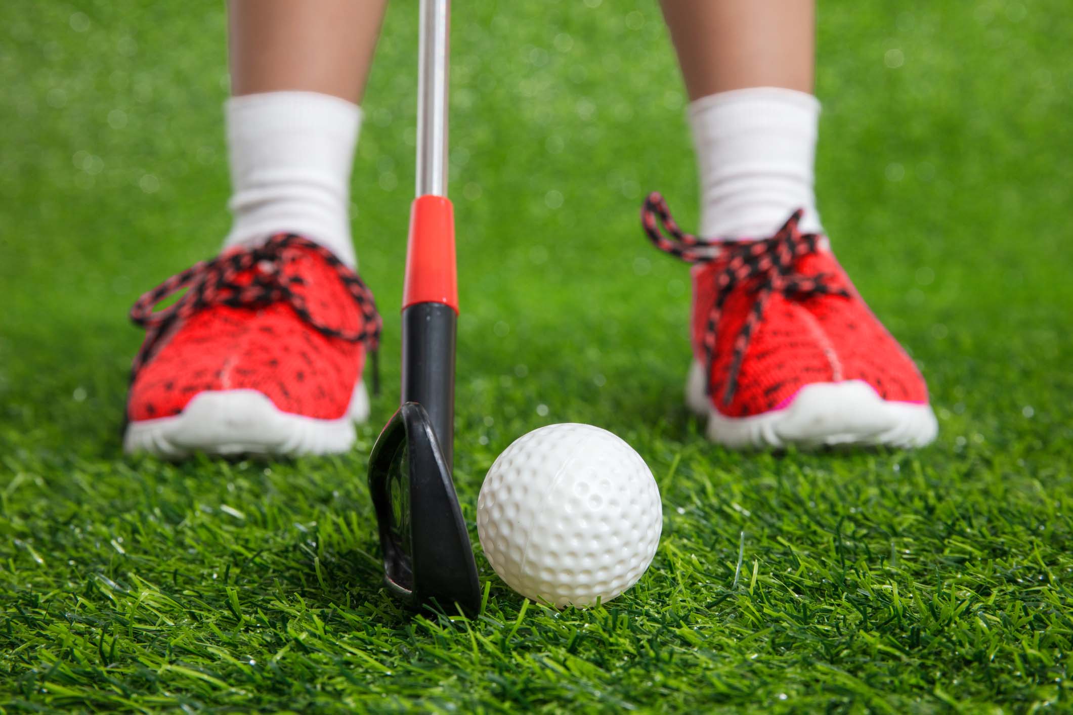Detail shot of a young person lower legs and a close up of the red sport shoes and white socks with a golf club and ball on grass